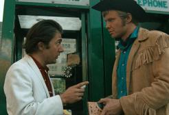 Shot from Midnight Cowboy movie, man in white suit faces man dressed as a cowboy in front of a phone box. 