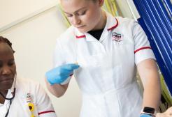 Two female nurses tending to a patient, one is listening to a stethoscope and the other is checking her watch