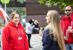 Three student ambassadors, in red University-branded hoodies, chatting to member of staff with concrete University building in the background