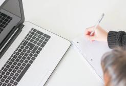 Person working at a white desk with laptop computer in front of them, writing notes on a notepad