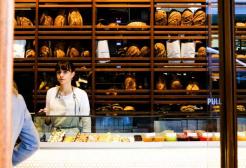 Bakery worker stands behind counter with shelves of bread behind her