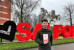 Student holding a book standing in front of the Salford sign, University of Salford