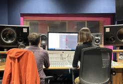 A man and a woman sit at a mixing desk in a recording studio