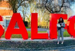 A female graduate holding a bunch of flowers stood in front of the Salford sign on Peel Park campus