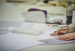 A student’s hands sketching on a desk, with designs on paper laid out around her
