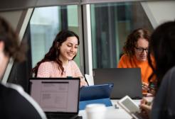 Students engaged in a group study session, smiling and working on laptops and tablets in a modern classroom setting.