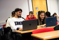University students sitting at desks in a classroom, smiling and working on laptops