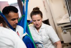 Male and female Occupational Therapy students practising moving and handling techniques with a sling and hoist under the direction of a tutor.