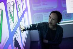A student interacts with a large digital screen displaying a grid of icons with checkmarks, representing completed tasks. The room, part of a digital immersive environment (DIVE), allows students to engage directly with the projected content, enhancing the learning experience.