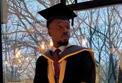 International student Ikenna in graduation robes stood in front of a window looking to the side