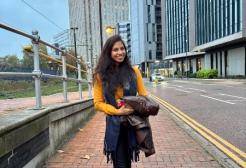 A female student on the pavement at the side of the road smiling at the camera holding a black coat in her arms