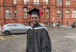 Femi Olayiwola in cap and gown standing in front of Peel Building, University of Salford
