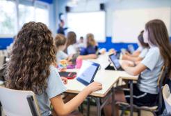 Children are shown using ipads in a classroom environment. They are facing away from camera towards the teacher at the front of the room.
