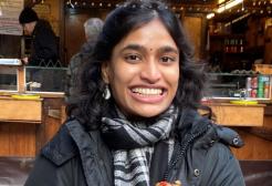 A female student in a coat and scarf smiling directly at the camera outside a Christmas Markets hut