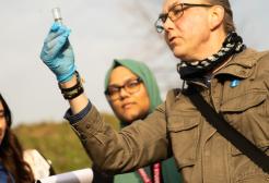 A lecturer and two students on a geography field trip. The male lecturer is holding up a test tube and looking directly at it. Two female students are stood behind him looking up at what is happening in the test tube.