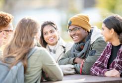 Group of male and female students sat around a table chatting outside