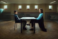 Characters sit across a desk from one another in a police holding centre
