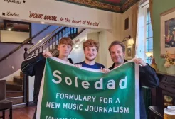 Three men stand in a pub holding a banner which reads soledad