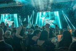 An award ceremony is taking place. People are sat around tables under blue sparkly lighting. A screen at the front of the room reads "Student Nursing Times Awards"