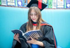 Female Graduate in Graduation cap and gown sits and reads the Graduation Brochure which is navy blue with silver foil diamond patterns