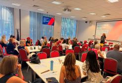 A presenter stands at the front of a busy room of event attendees. The presenter, Professor Jane McAdam, smiles as she addresses the room.