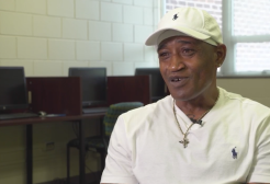 A man in a white shirt and white baseball cap speaks to a camera