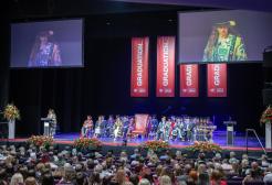 A crown can be seen watching a Graduation ceremony. A procession sits on stage in various gowns. Red banners reading graduation can been seen hanging in the back of the stage. 