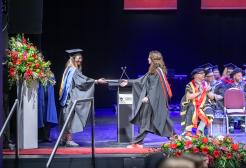 A graduate in a black gown with a red, grey and dark blue hood, crosses the stage at their Graduation. The Graduate holds out their hand to receive a black scroll with gold detailing on it. The scroll is handed to them by a member of staff wearing a black gown with a yellow and blue hood and mortar board hat. A procession sits on stage behind the pair and applauds. A large bouquet of flowers can be seen at the end of the stage on a white pillar.