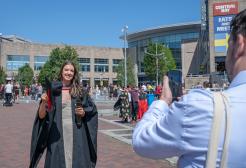 A graduate smiles as their picture is taken using a phone, which can be seen being held in the corner of the picture. The graduate holds a black mortar board hat with a red tassel and wears a black graduation gown on top of a long stone coloured dress.