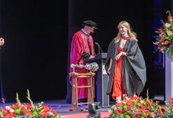 A graduate in a long red dress and black gown smiles as they walk across the stage. They walk towards the presiding officer who can be seen from the back wearing a blue and gold gown. In the background the presenter can be seen wearing a red gown.