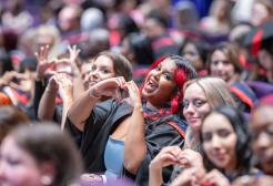 A graduate in the theatre audience makes a heart sign with her hands and smiles at the camera.