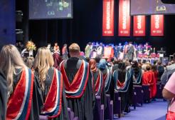 Graduates are photographed from behind stood at the end of their assigned rows in the Lowry Theatre. The graduates wear black graduation gowns with hoods on top. The hoods are dark blue and grey on the outside, and red in the middle with the University of Salford logo repeated over the red in black.
