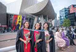three graduates in Graduation gowns hold bouquets of red flowers and smile at the camera while standing in front of the Lowry theatre.