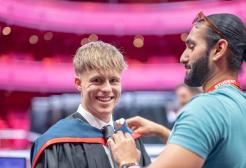 Graduate smiles at the camera as a student ambassador helps them put their graduation gown over their suit. The gown is black and the hood is red and dark blue.