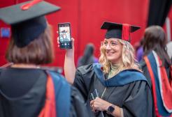 A graduate in a black graduation gown and blue and red hood, holds up their phone for another graduate stood across from them in the same graduation attire. Both wear black mortar board hats with red tassels