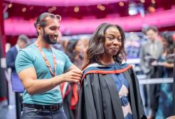 A graduate smiles at the camera while wearing a black gown over their blue and white dress. A student ambassador adjusts their graduation hood which is blue and red from behind them.