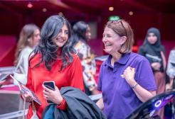 A graduate in a red dress holds a Graduation brochure, graduation ticket and their black robe as they laugh with a staff member in a purple polo shirt.