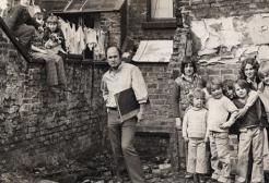 photograph of a group of children and an adult man standing in a rundown urban courtyard with brick buildings and broken windows in the background, suggesting poor housing conditions.