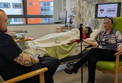 A man and a woman sat in a hospital simulation suite, filming a podcast with a simulation dummy in the background