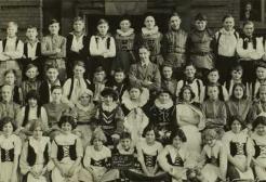 Black-and-white group photograph of approximately 60 schoolchildren and several adults posed in four rows outside a brick building. Many of the children wear coordinated costumes with dark vests or bodices over light shirts, suggesting a school performance or class production.