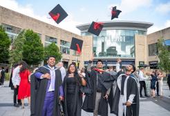 Graduates wearing caps and gowns celebrate outdoors, they smile at the camera and throw their caps into the air above a crowd of people. Trees surround the area and the sky is partly cloudy.