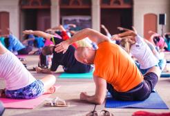 A diverse group of people sit on yoga mats facing away from the camera. They are all leaning to one side, with one arm raised over their heads as they stretch and reach sideways.