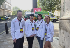 A group of four podiatry students and staff smile at the camera. They are stood on the street in front of a number of support vehicles and wear official London Marathon branded jackets and lanyards.