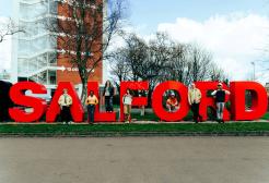 A group of six people pose in front of the Salford sign