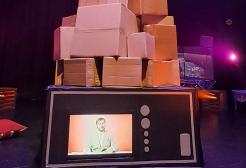 Stage production set featuring an oversized prop television displaying a person talking, topped with a stack of cardboard boxes