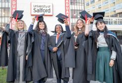 Five graduates in academic gowns stand outside a University of Salford building, holding their mortarboards above their heads. One graduate holds a rolled certificate while the group poses in front of a large sign reading “University of Salford.” Modern campus buildings and graduation banners can be seen in the background.