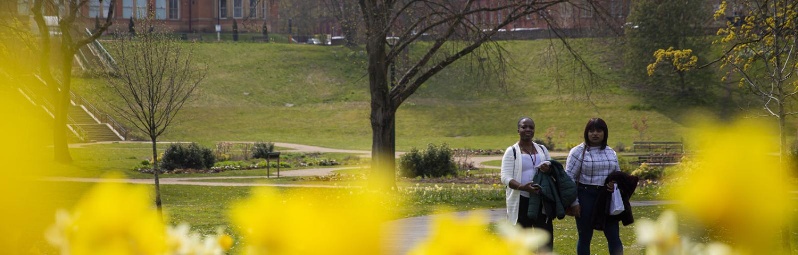 Two students walking through Peel Park in spring with daffodils in the foreground