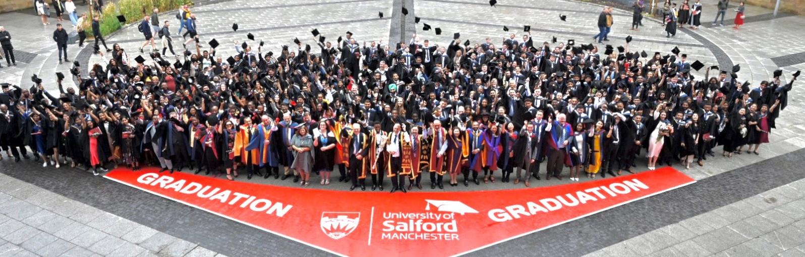 Large group of people posing for a photograph in graduation attire in front of floor sign that reads 'Salford University Manchester Graduation'. 