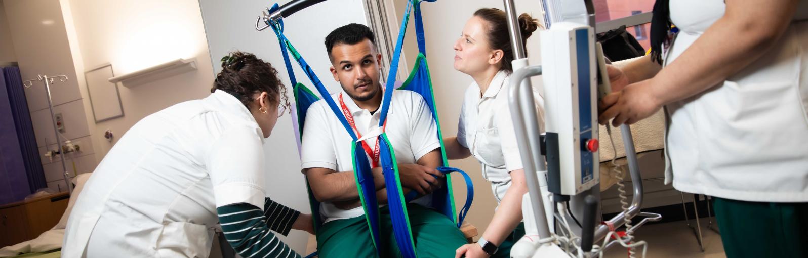 Male and female Occupational Therapy students practising moving and handling techniques with a sling and hoist under the direction of a tutor.