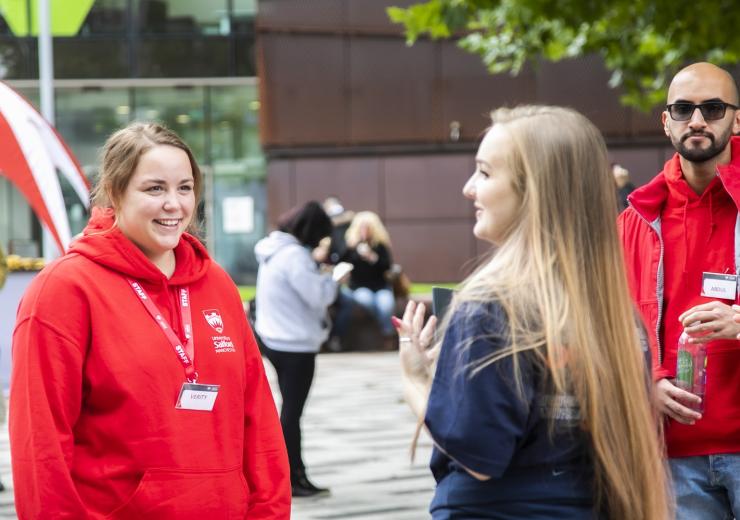 Three student ambassadors, in red University-branded hoodies, chatting to member of staff with concrete University building in the background